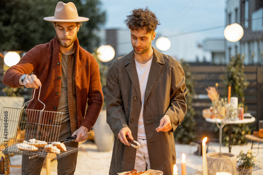 Two male friends serving grilled sausages on a dining table outdoors. Fun summer time in a country h