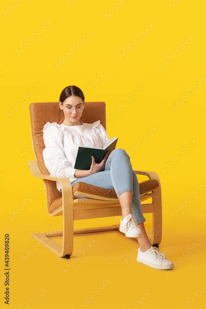 Young woman reading book while sitting in armchair on color background