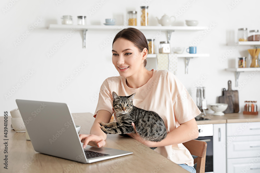 Beautiful young woman with cute cat using laptop in kitchen