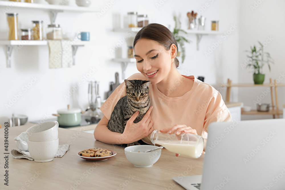 漂亮的年轻女人和可爱的猫在厨房把牛奶倒进碗里