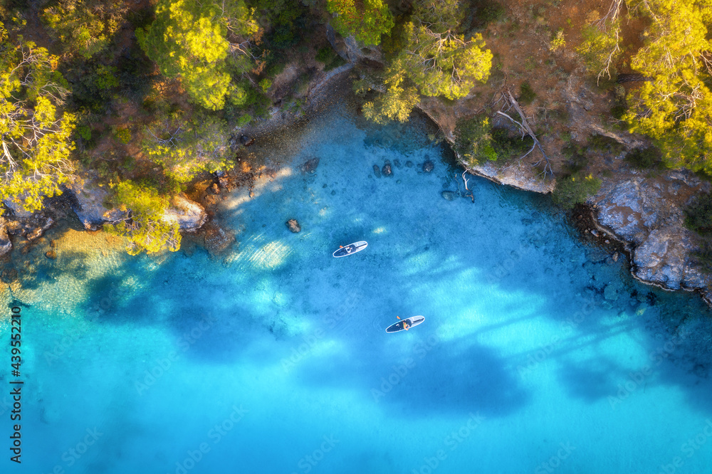 Aerial view of people on floating sup boards on blue sea, rocks, trees at sunset in summer. Blue lag