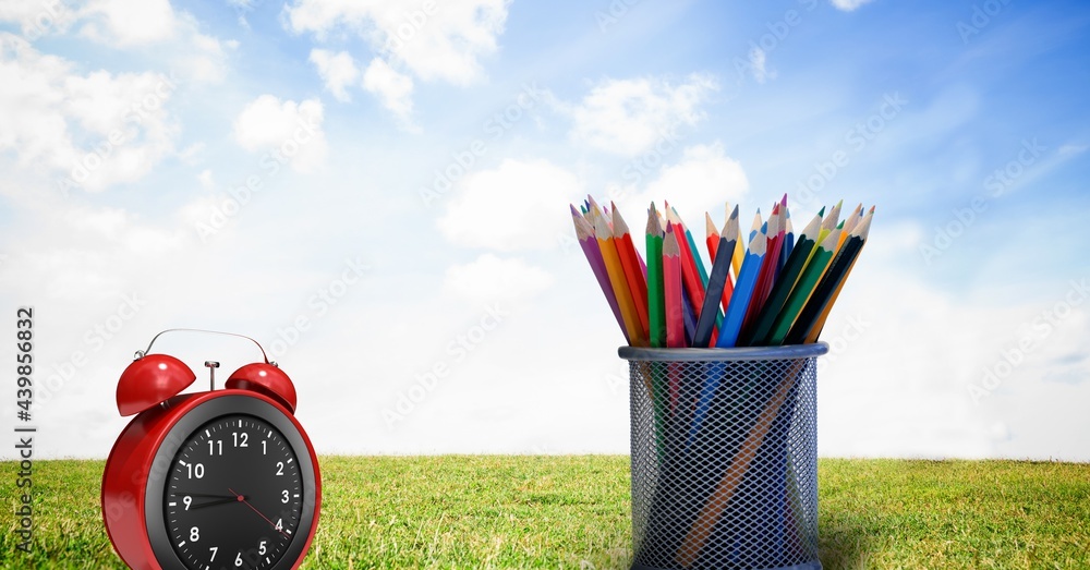 Red alarm clock and multiple colorful pencils in stand against landscape with grass and blue sky