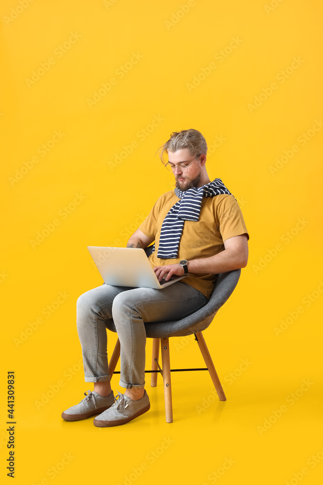 Young man with laptop sitting in armchair on color background