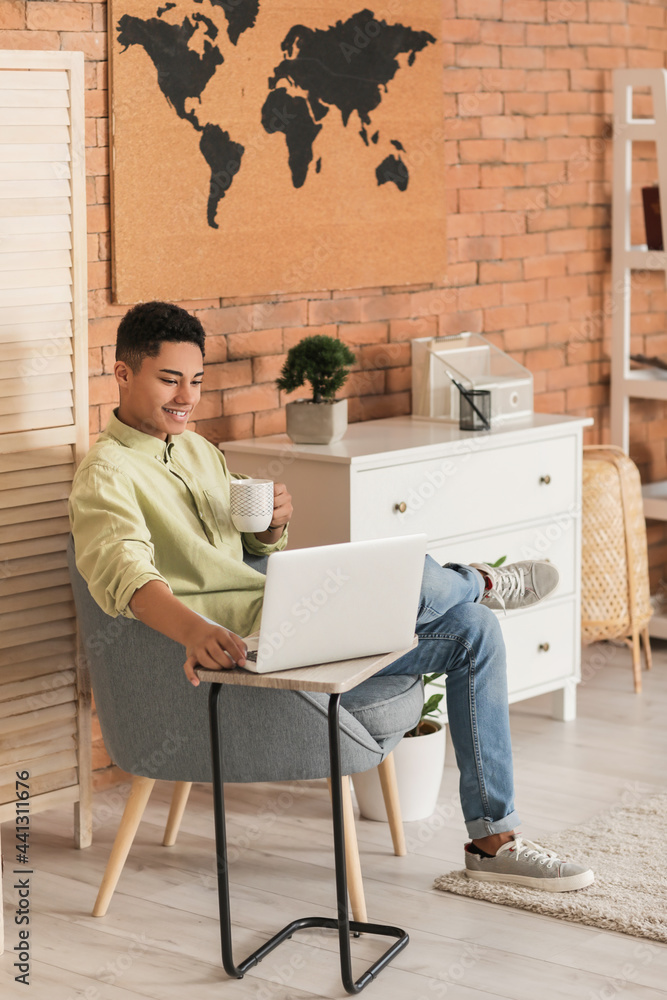 Young African-American guy with laptop drinking coffee while sitting in armchair at home