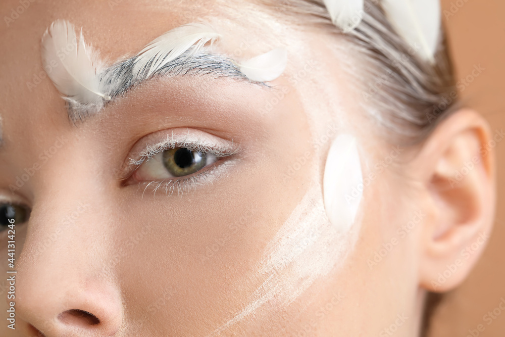 Beautiful young woman with dyed eyebrows and feathers on color background, closeup