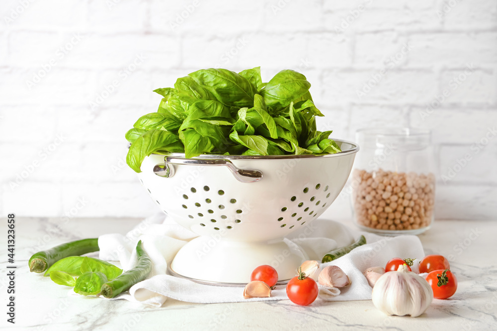 Colander with fresh basil leaves and different products on table against brick wall