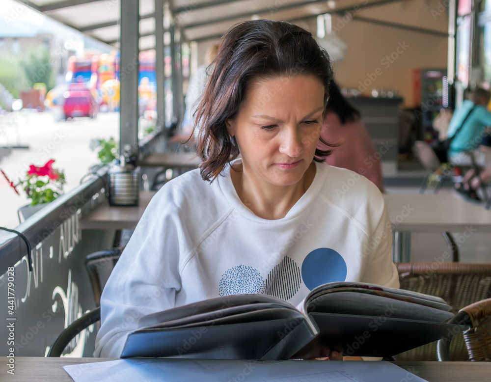 Caucasian middle-aged woman is choosing a dish at a menu in a street cafe.