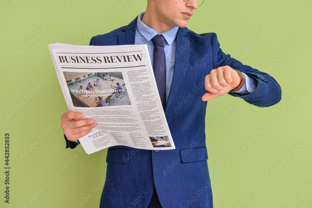 Young businessman with newspaper looking at wristwatch on color background