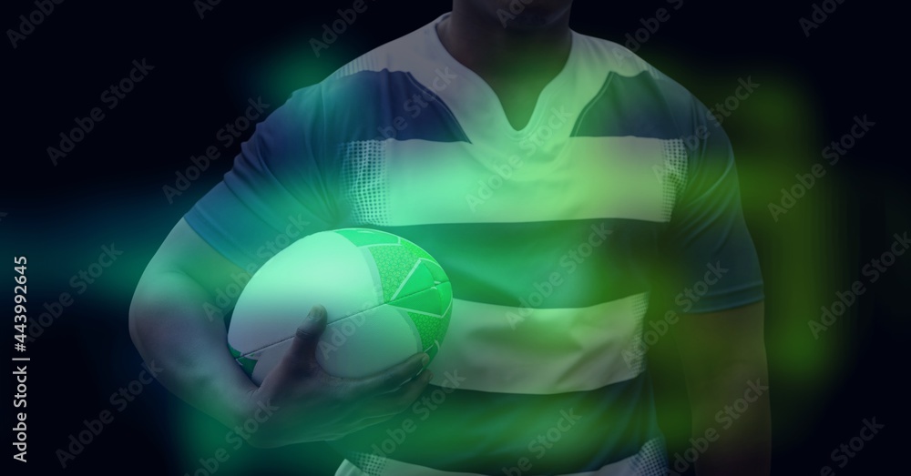 Composition of glowing green blur over male rugby player holding rugby ball, on black