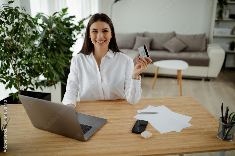 Young smiling woman holding credit card and using laptop computer. Businesswoman working at home.