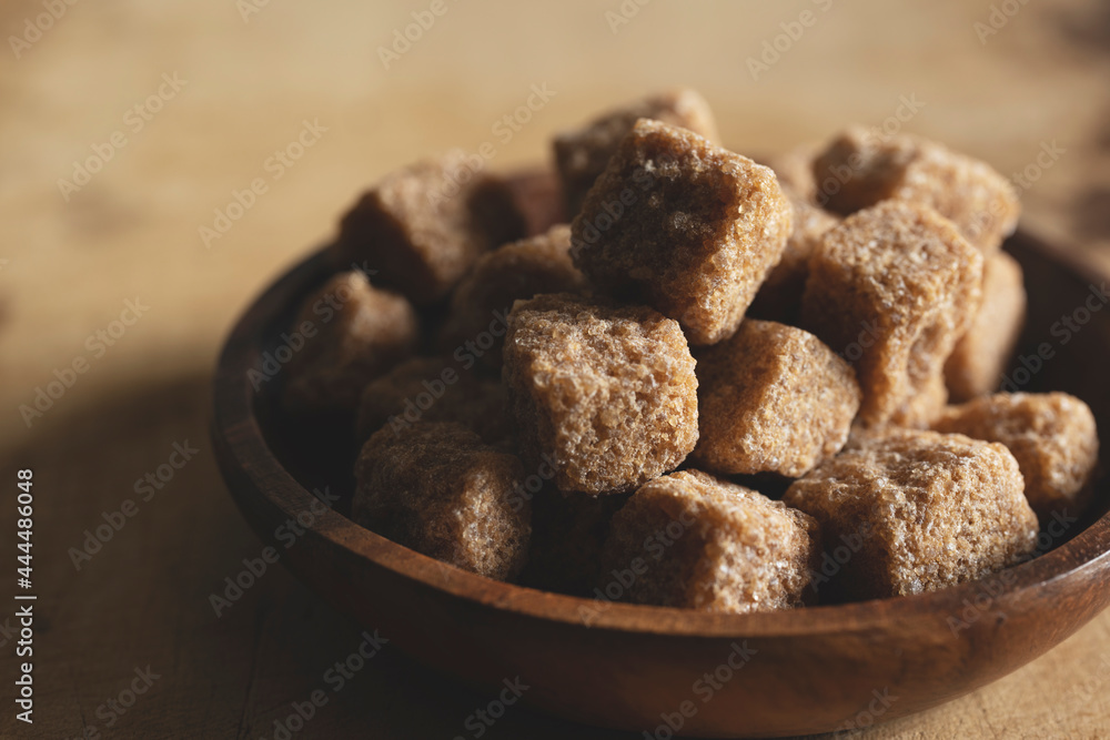 Brown cane sugar cubes in a wooden dish set against a wooden background.