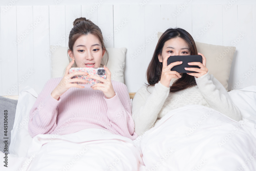 Two beautiful women lying on the bedside playing mobile games