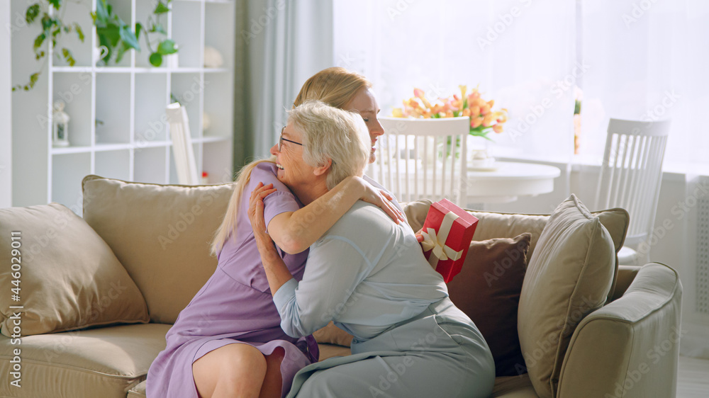 Long haired blonde in purple dress gives small box with present to aged lady with joyful smile