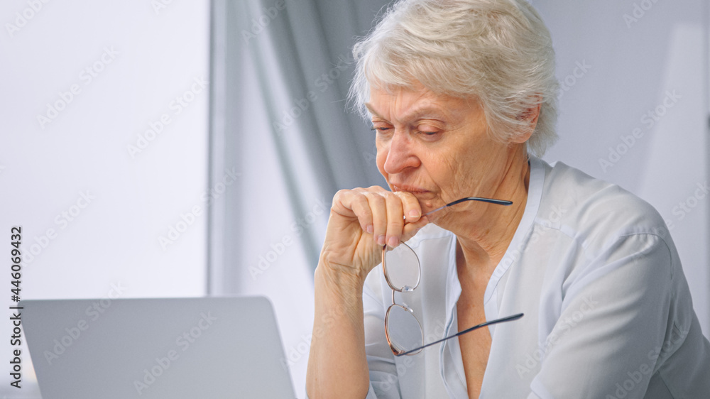 Corporate lady manager pensioner takes glasses off relaxing from work and sitting at grey laptop aga