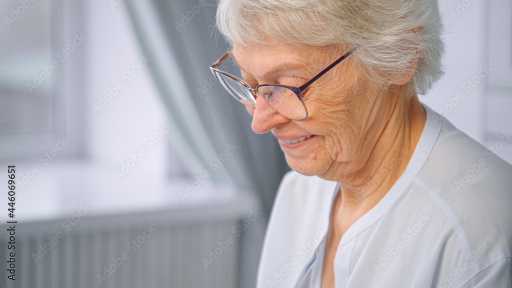 Delighted senior woman with short grey hair and glasses types on white smartphone and smiles against