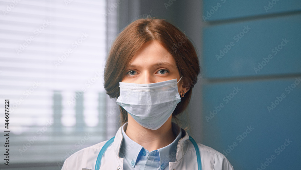 Red haired woman with face mask and looks straight standing in local hospital room