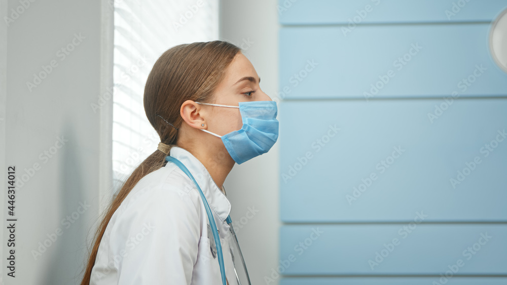 Exhausted young woman in white coat with stethoscope on neck takes off blue disposable face mask