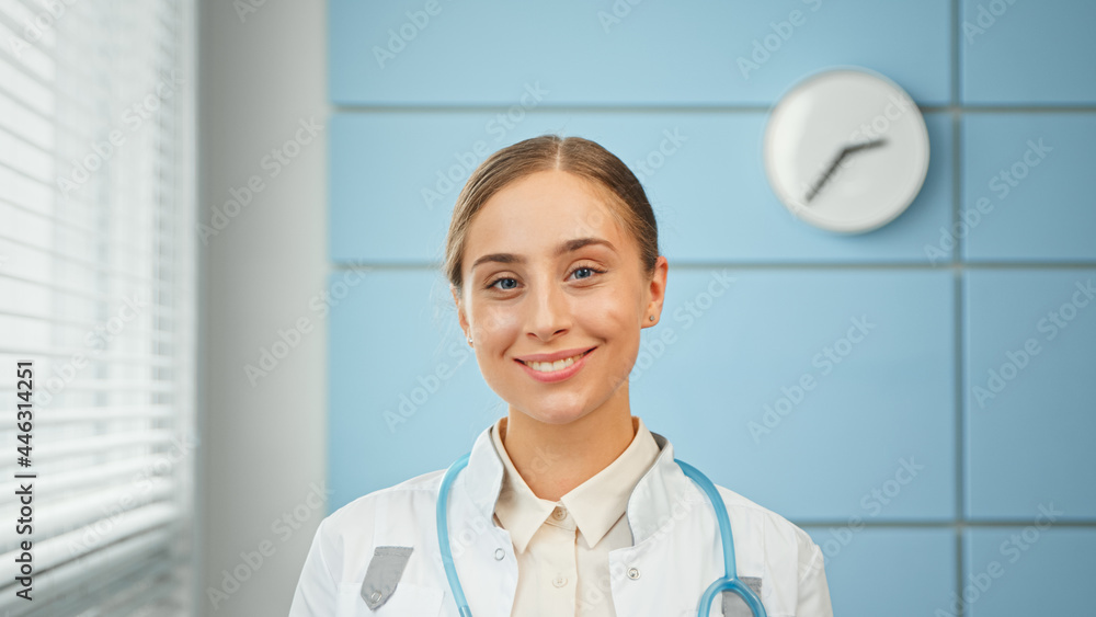 Blonde young woman general practitioner in white coat with blue stethoscope looks straight standing