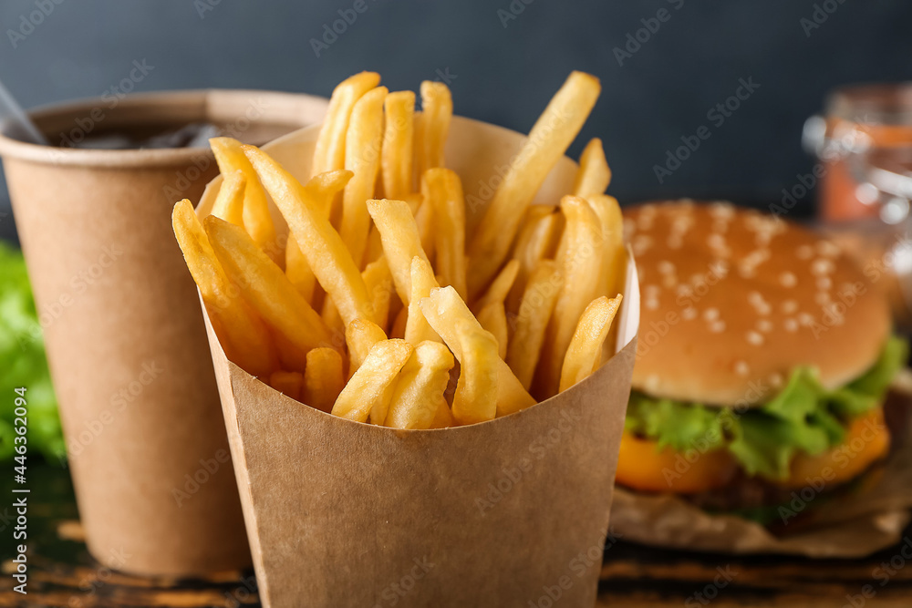 Paper box with tasty french fries, burger and cup of beverage on dark wooden background, closeup