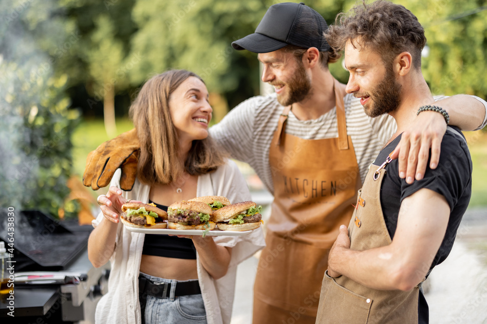 Young people enjoy yummy burgers made on a grill at picnic, standing together and having fun. Friend