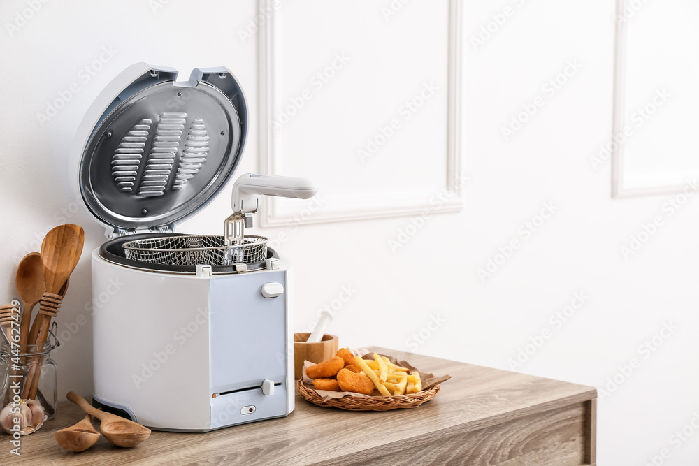 Modern deep fryer, french fries, nuggets and kitchen utensils on table near light wall