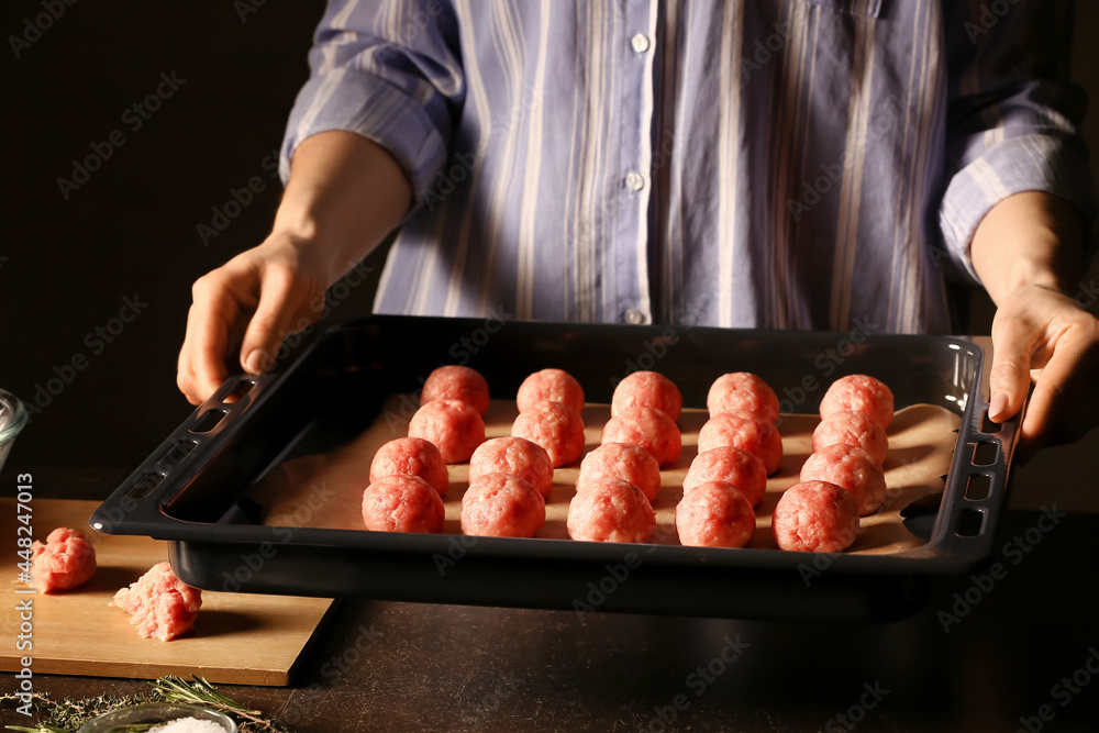 Woman holding baking tray with raw cutlets made of fresh forcemeat on dark background, closeup