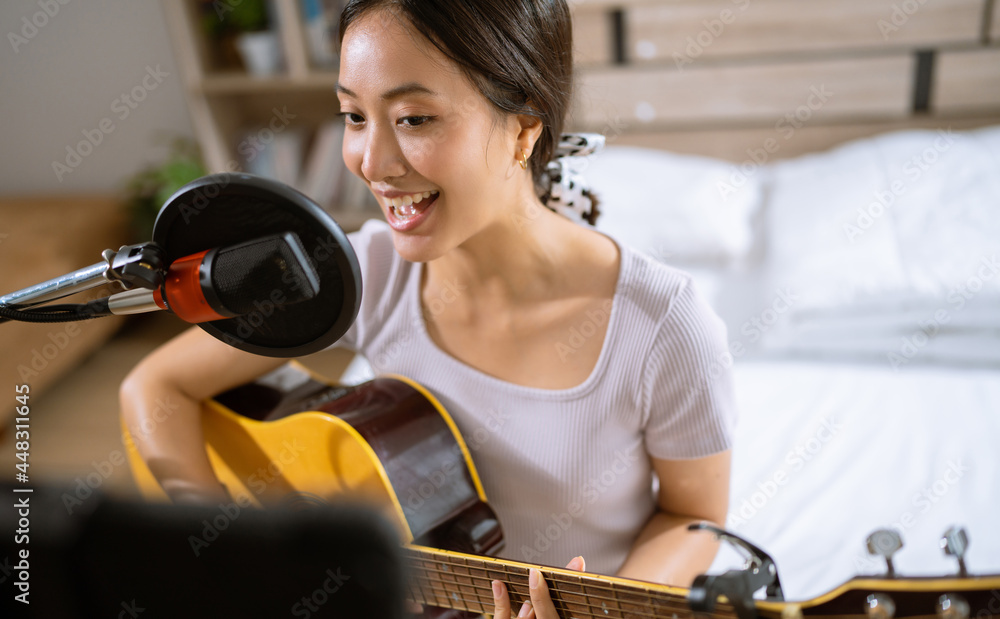 亚洲女人在卧室里弹吉他并录制人声。她是现场直播的。