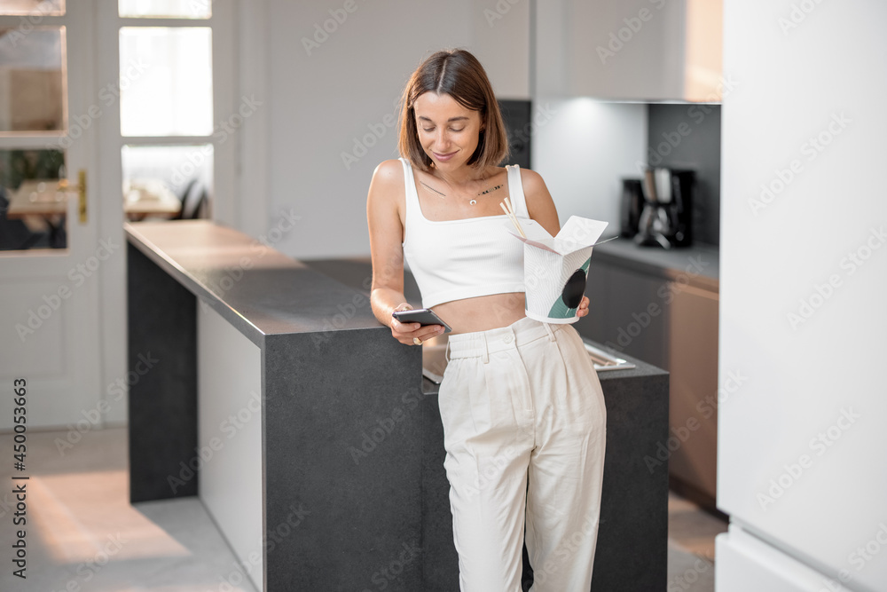 Portrait of a young woman with phone and takeaway asian food on the kitchen at home. Modern and heal