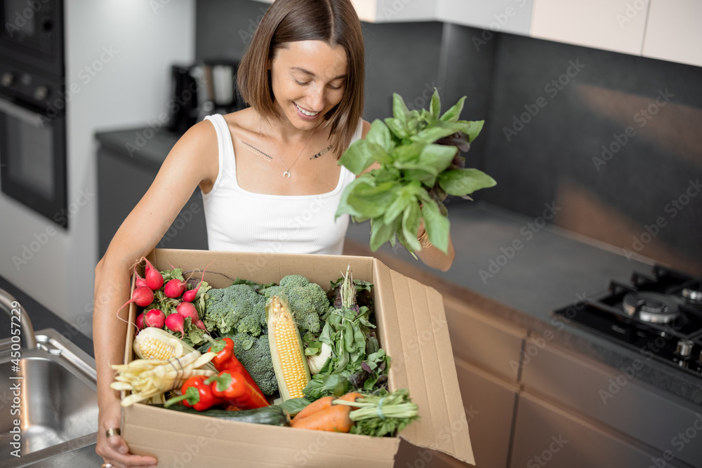 Young woman with fresh vegetables packed in cardboard box at kitchen. Delivering and buying food onl