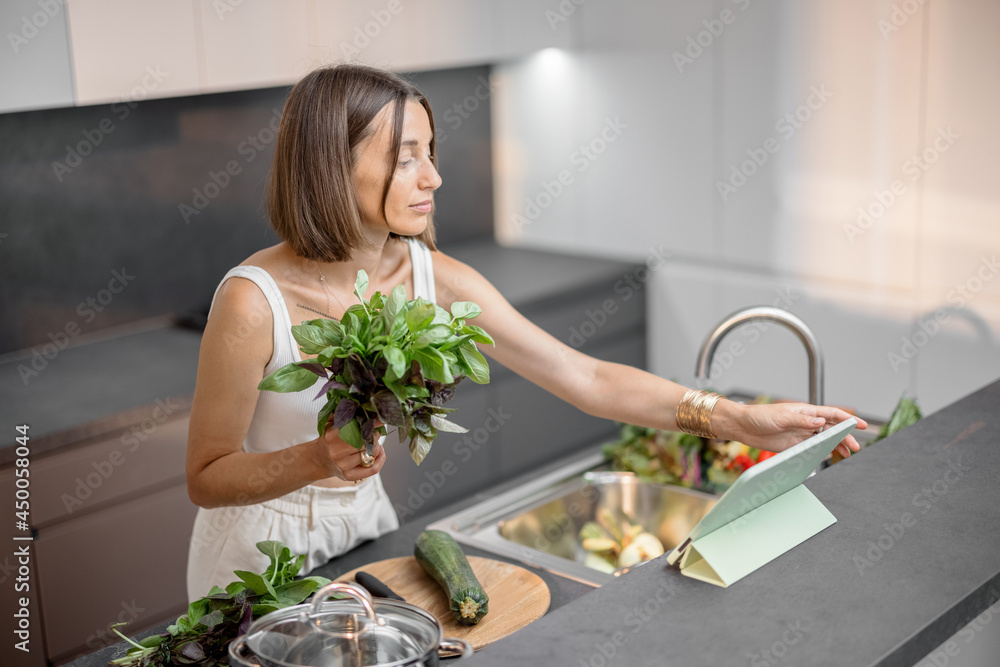 Young woman cooking with fresh vegetables and greens looking at recipe on the digital tablet at mode