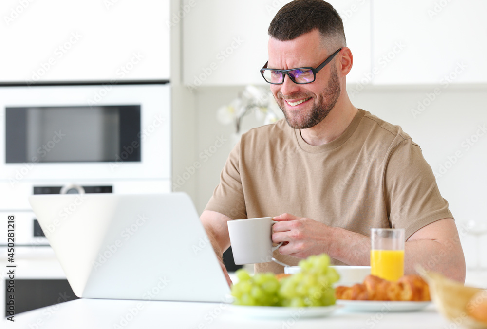 Young smiling handsome man working remotely on laptop in modern kitchen at home