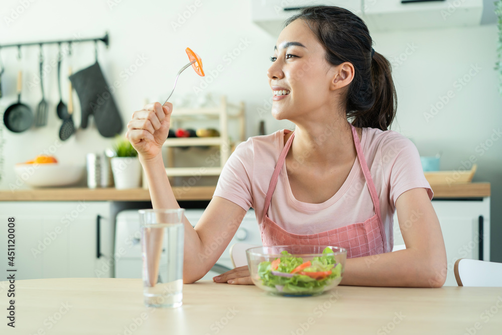 Asian attractive woman in apron eating green salad in kitchen at home. 