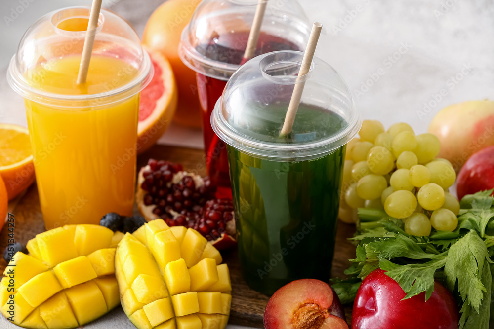 Plastic cups with healthy juice, fruits and vegetables on light background