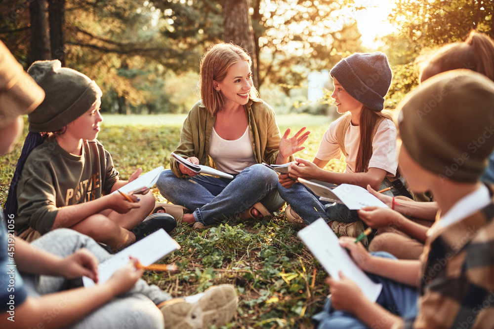 Children and smiling female teacher sitting in circle on grass in forest and talking about nature