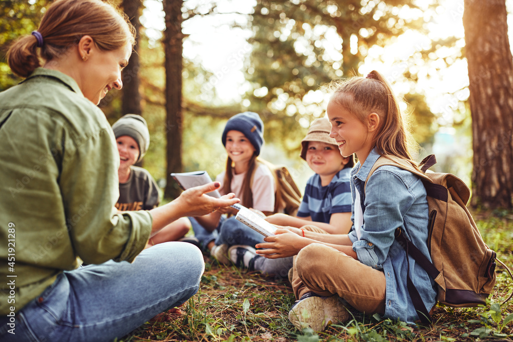 Children and smiling female teacher sitting in circle on grass in forest and talking about nature