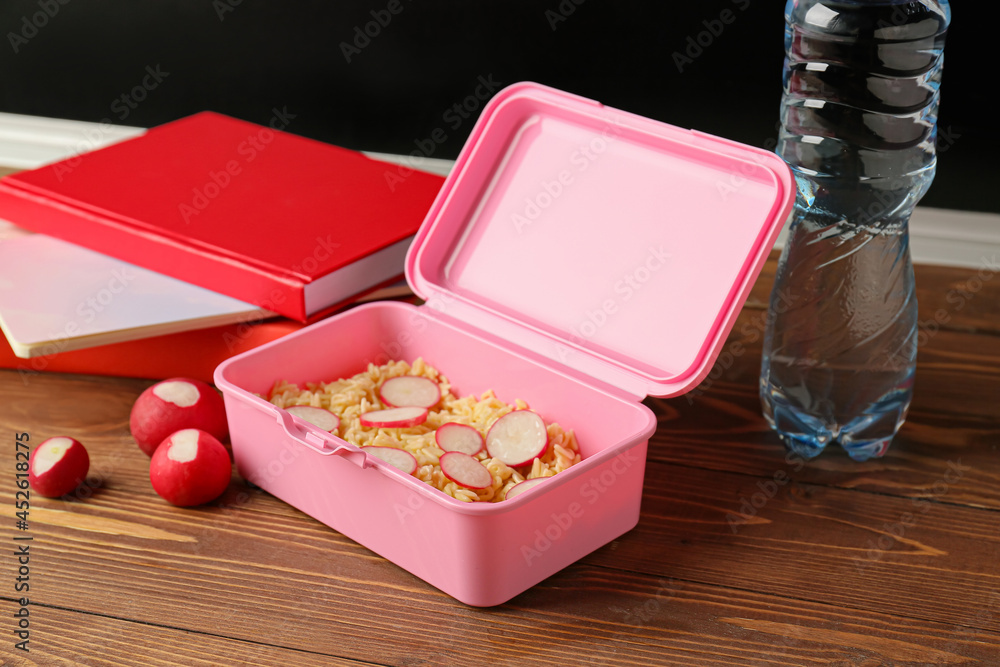 Lunch box with food, radish, bottle of water and notebooks on wooden table in classroom