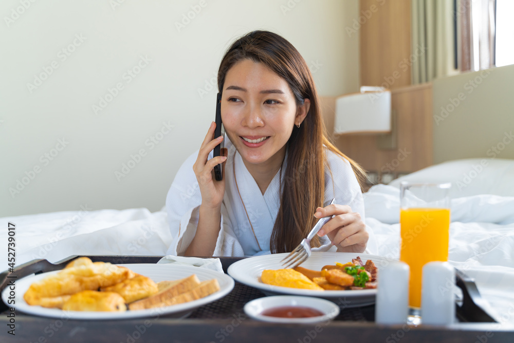 Portrait beautiful young Asian woman wearing bathrobe talking on a phone while eating breakfast on a