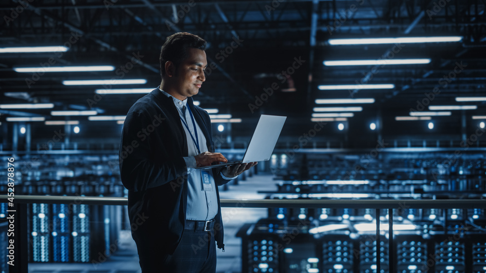 Portrait of IT Specialist Uses Laptop in Data Center. Server Farm Cloud Computing Facility with Male