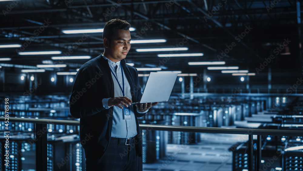 Portrait of IT Specialist Uses Laptop in Data Center. Server Farm Cloud Computing Facility with Male