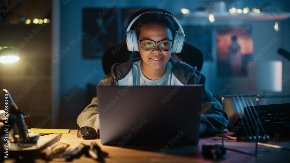 Young Teenage Multiethnic Girl Using Laptop Computer and Wearing Headphones in a Dark Cozy Room at H