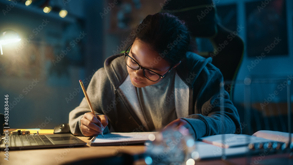 Young Teenage Multiethnic Black Girl Writing Down Homework in a Notebook with a Pencil, Using Laptop