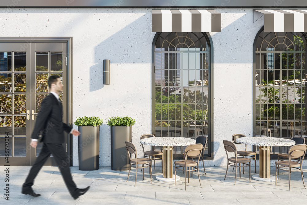 Young man in suit walking past creative concrete cafe exterior with terrace furniture in daylight.