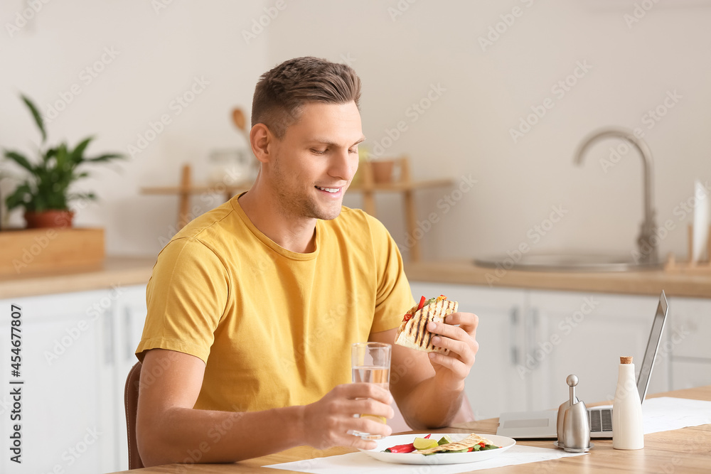 Handsome young man with laptop eating tasty quesadilla at home