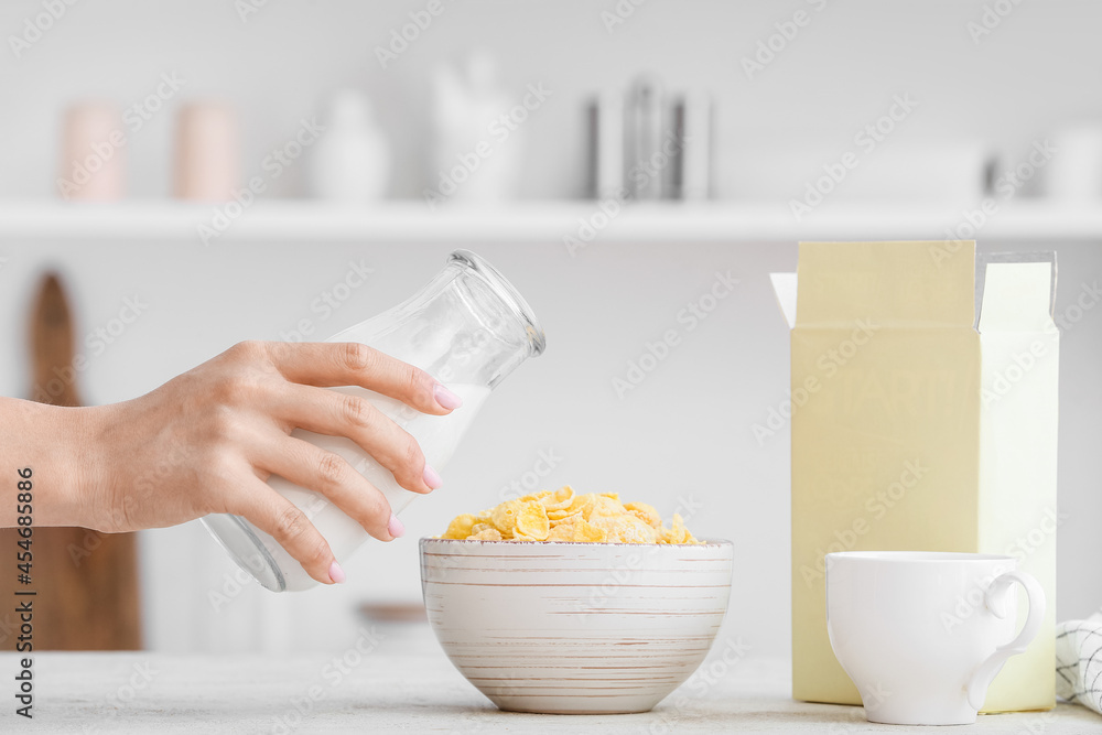 Woman pouring milk from bottle into bowl with corn flakes on table in kitchen