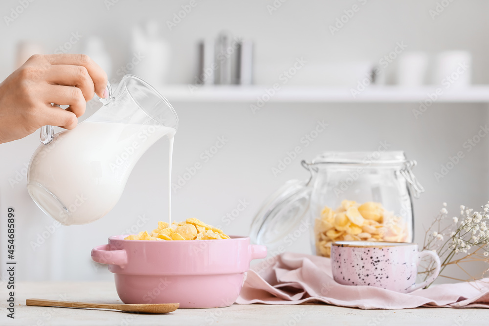 Woman pouring milk from jug into pot with corn flakes on table in kitchen