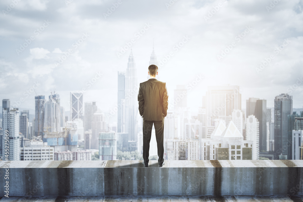 Back view of young businessman standing on concrete edge of building on city background with mock up