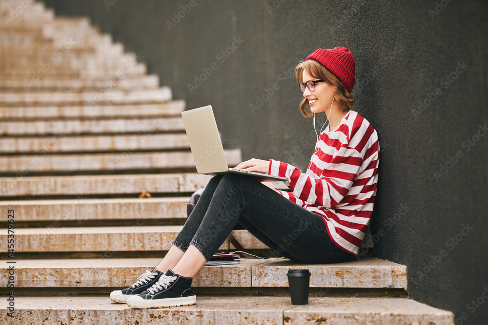 Student girl in earphones sits outside on staircase with laptop, notebooks and takeaway coffee