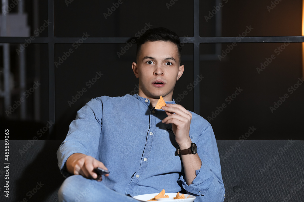 Handsome young man eating tasty nachos while watching TV at home late in evening