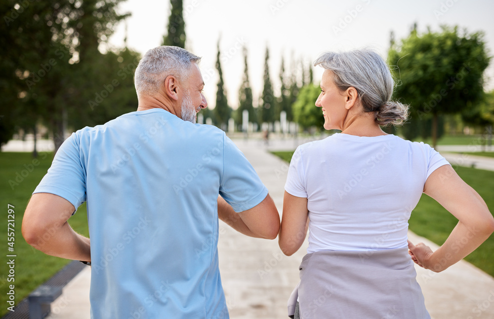 Photo from back of active mature couple smiling while jogging together in park in morning