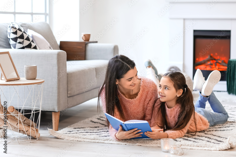 Young mother with daughter reading book near fireplace at home